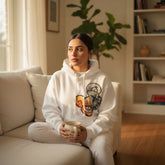 Person wearing a white hoodie with a graphic design, sitting on a couch holding a mug in a cozy living room.